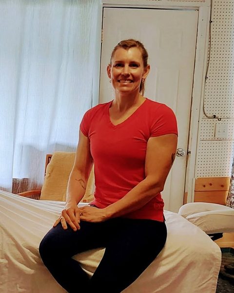 Jess, smiling, sitting on the massage table in her Wynndel studio