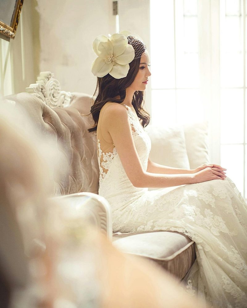 A bride in a lace gown seated by a tall window, looking out into the light